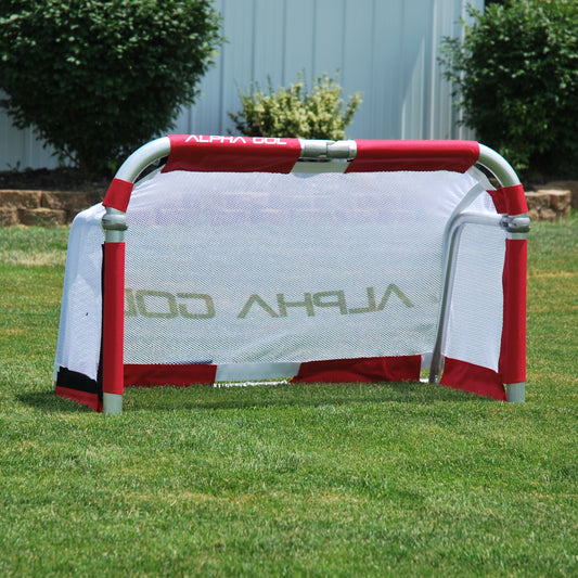 A small Alpha Gol soccer goal with a red and white frame and a white mesh back, set up on a green grass lawn with bushes and a white fence in the background.