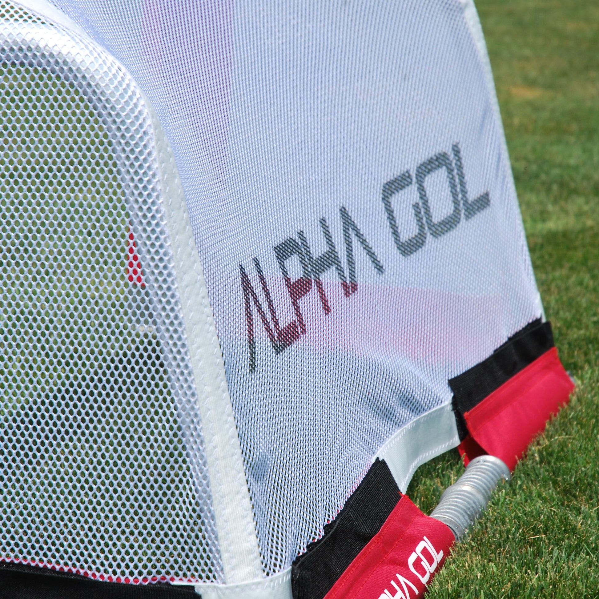 Close-up of a golf practice net on a grassy field, showing a white mesh back wall with the brand name ALPHA GOL printed in gray and red trim.