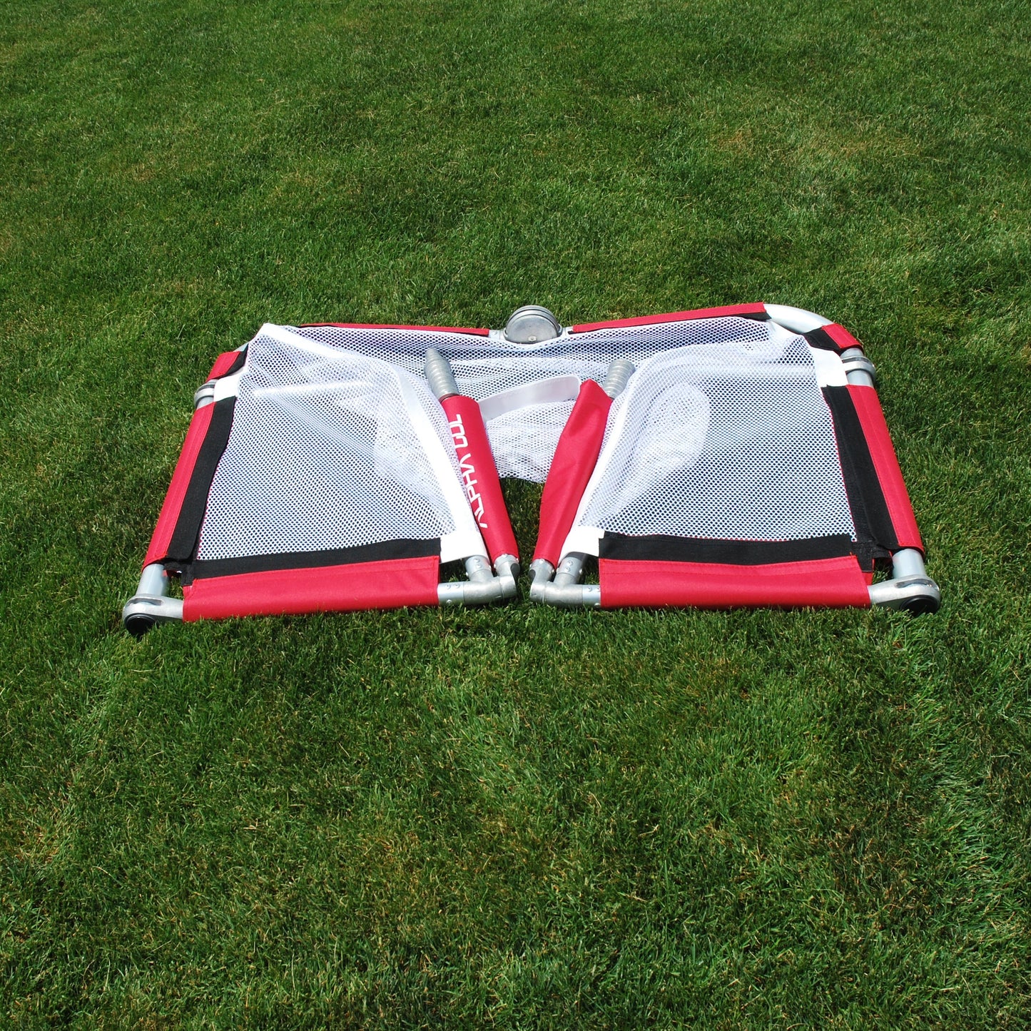 A red and white mesh foldable soccer goal laid flat on a green grass lawn.