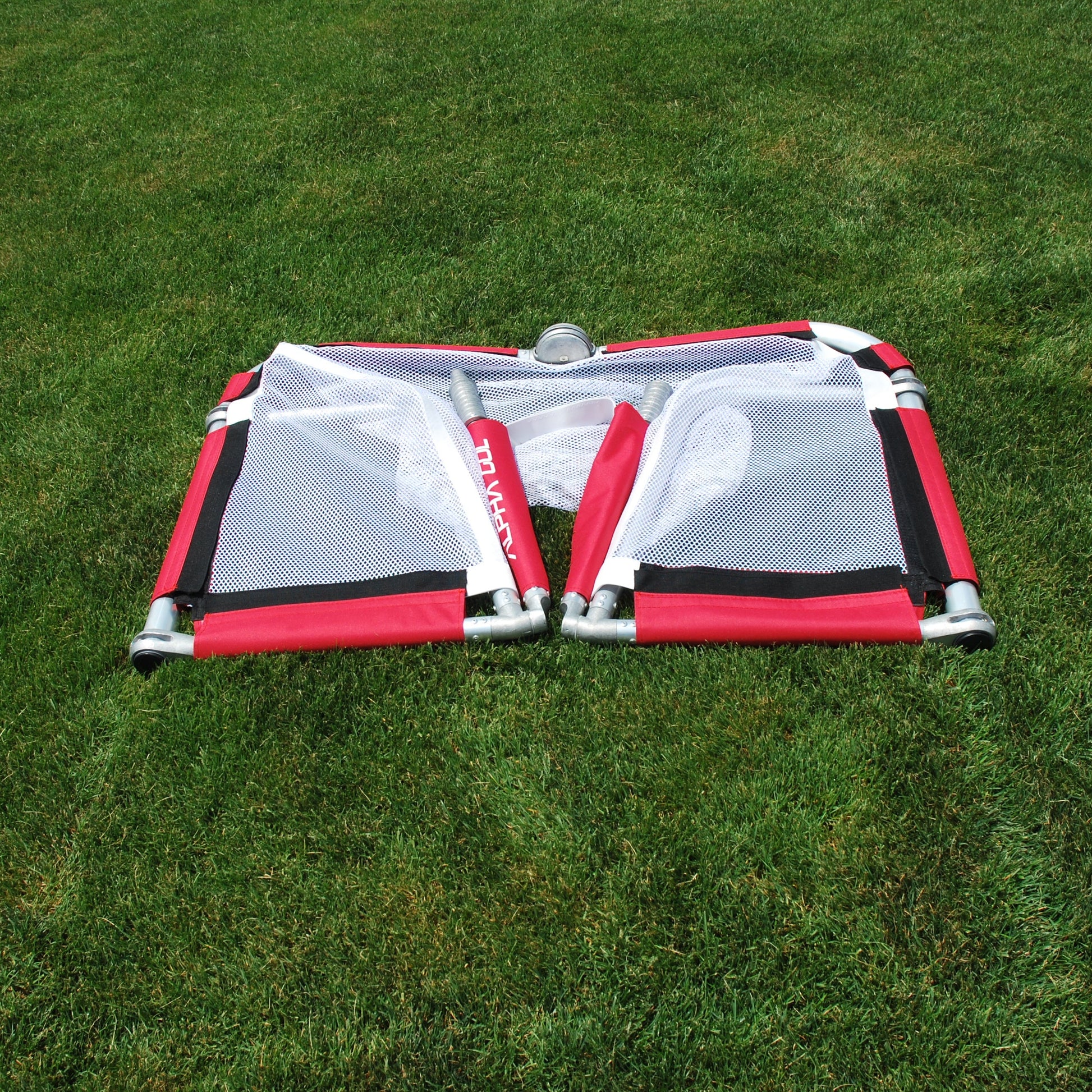 A red and white mesh foldable soccer goal laid flat on a green grass lawn.