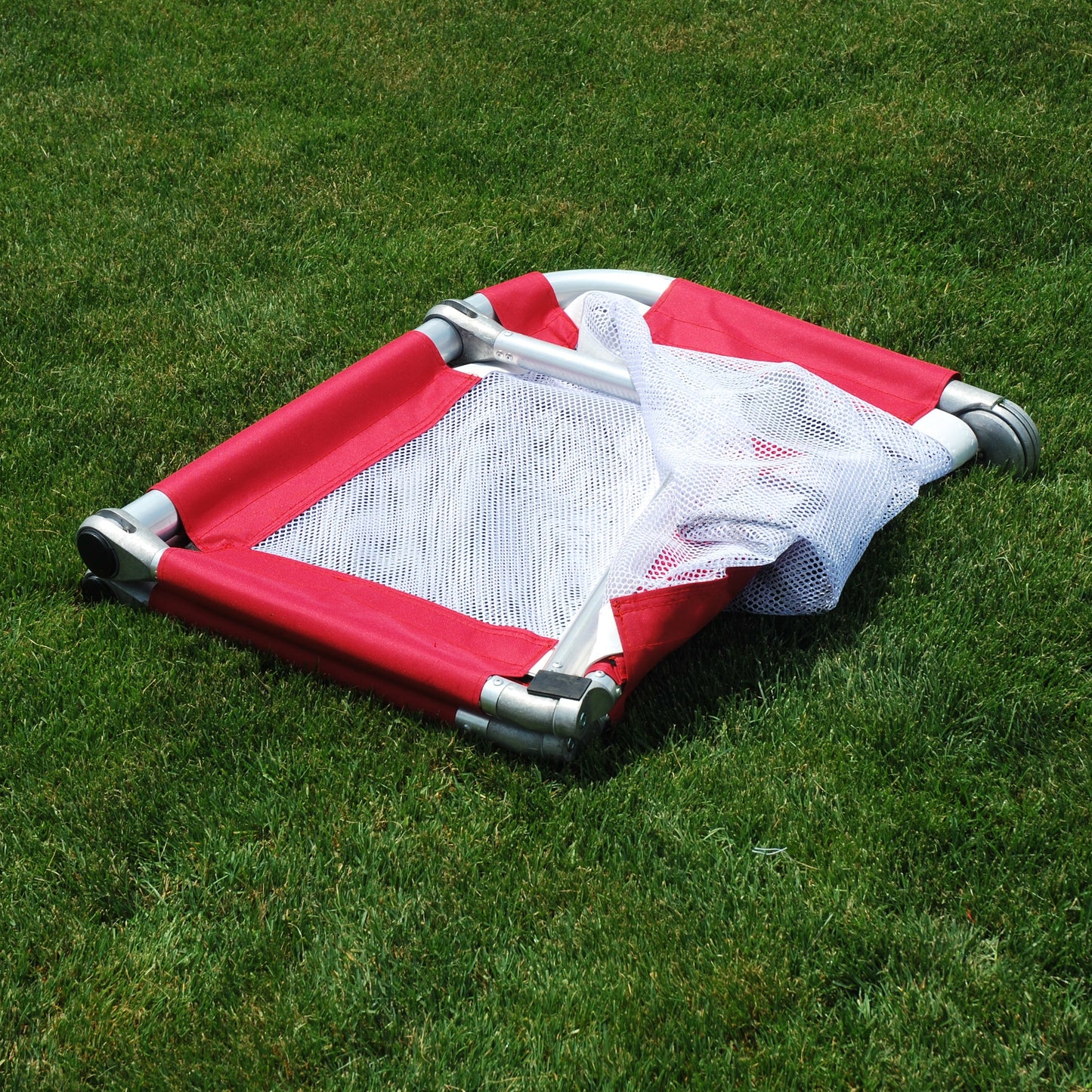 A red and white mesh soccer goal with gray metal corner supports, lying on a green grass lawn.